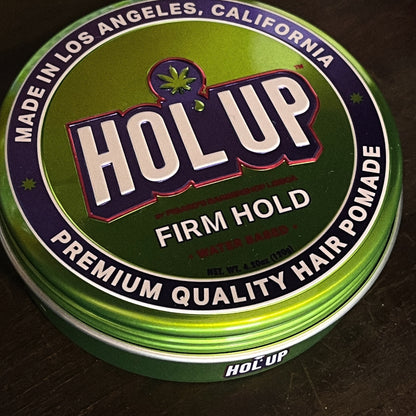 A round metal tin of Hol Up Firm Hold hair pomade, featuring a green and purple label with white lettering, photographed on a dark wooden surface.